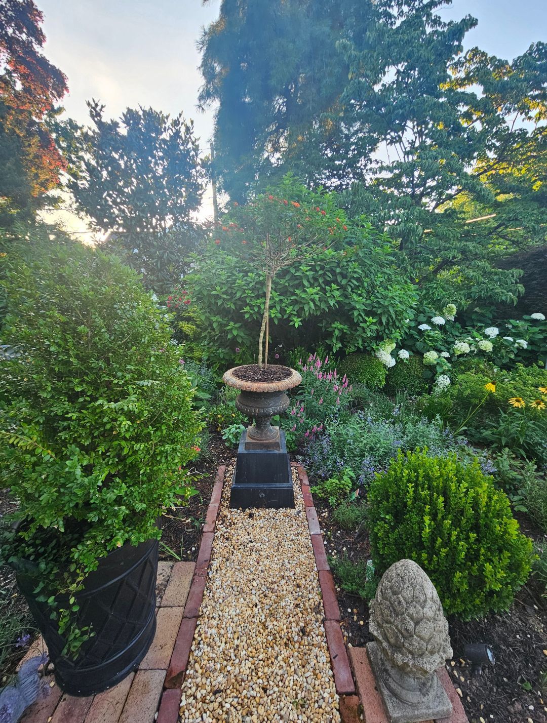 Narrow garden path with potted plants, hedges, and a birdbath in a lush backyard.