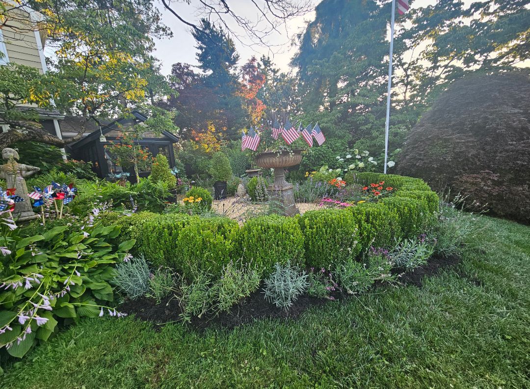 Lush garden with trimmed hedges, flowers, trees, and a small gazebo in warm sunlight