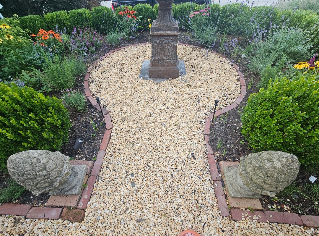 Stone garden monument centered on a gravel path, bordered by brick edging, rocks, and green shrubs.