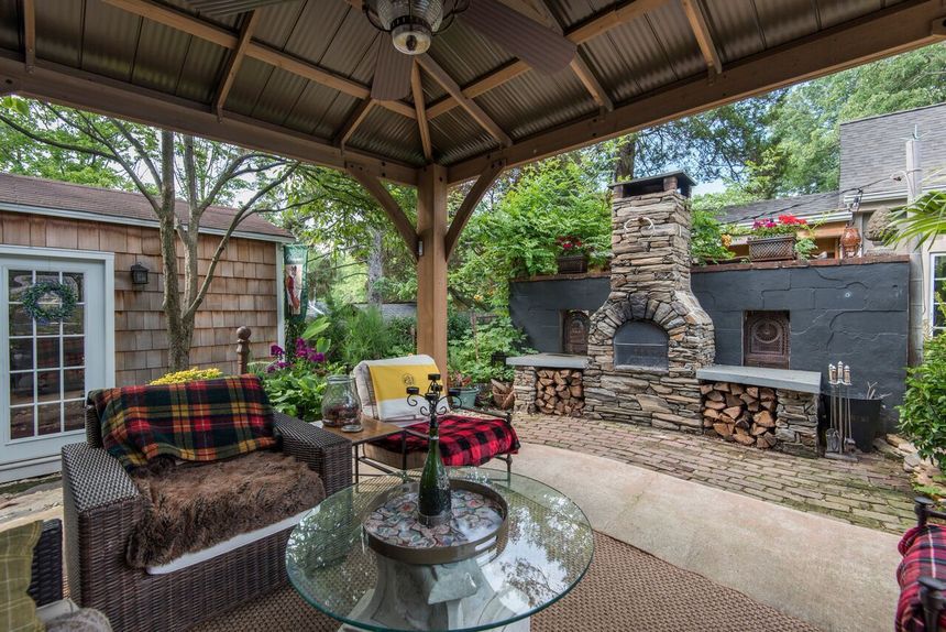 Covered patio with wicker seating, glass table, and stone fireplace in a backyard garden