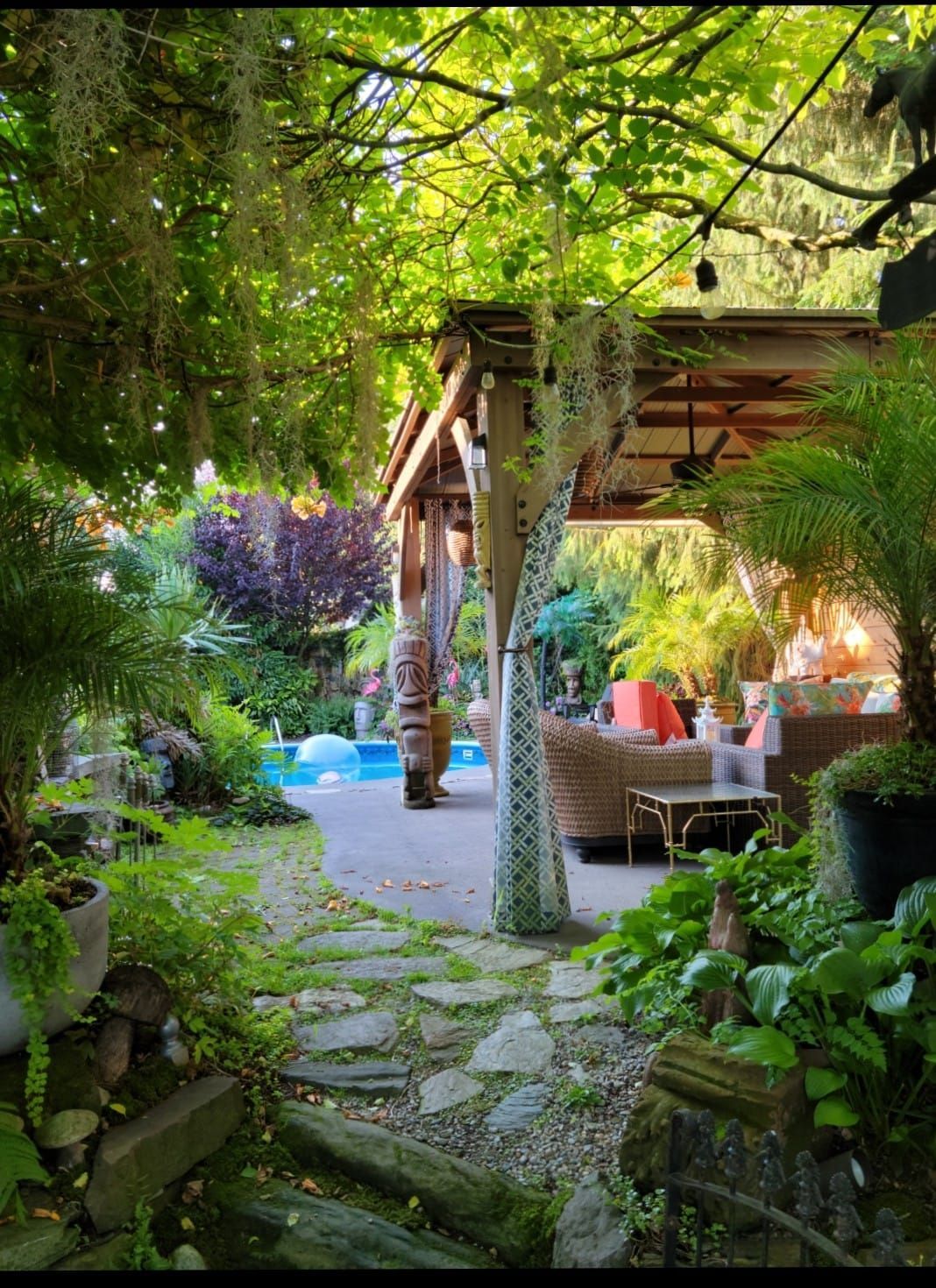 Lush garden patio with stone path, thatched pergola, wicker seating, and colorful flowers