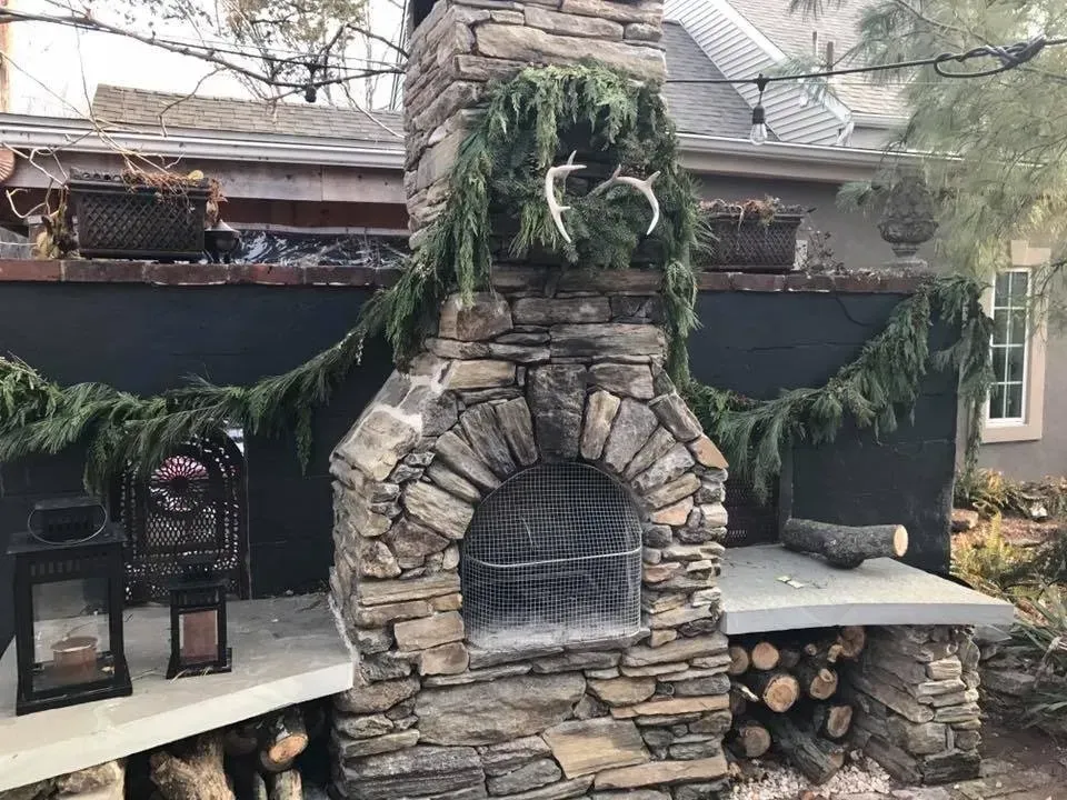 Stone outdoor fireplace with arched opening, evergreen garland, and benches with lanterns beside stacked firewood