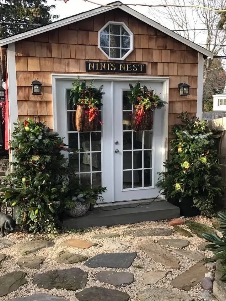Small rustic cottage storefront with white French doors, hanging plants, and a stone path.
