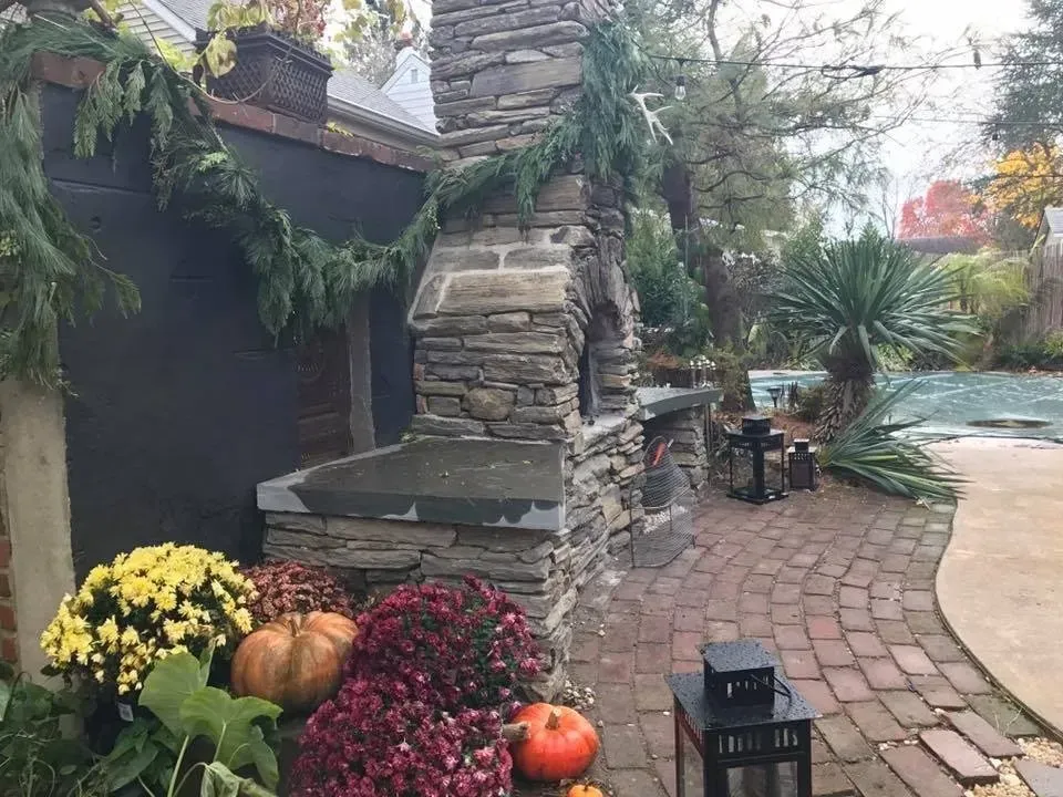 Brick patio with autumn pumpkins, yellow mums, and a stone outdoor fireplace in a garden courtyard