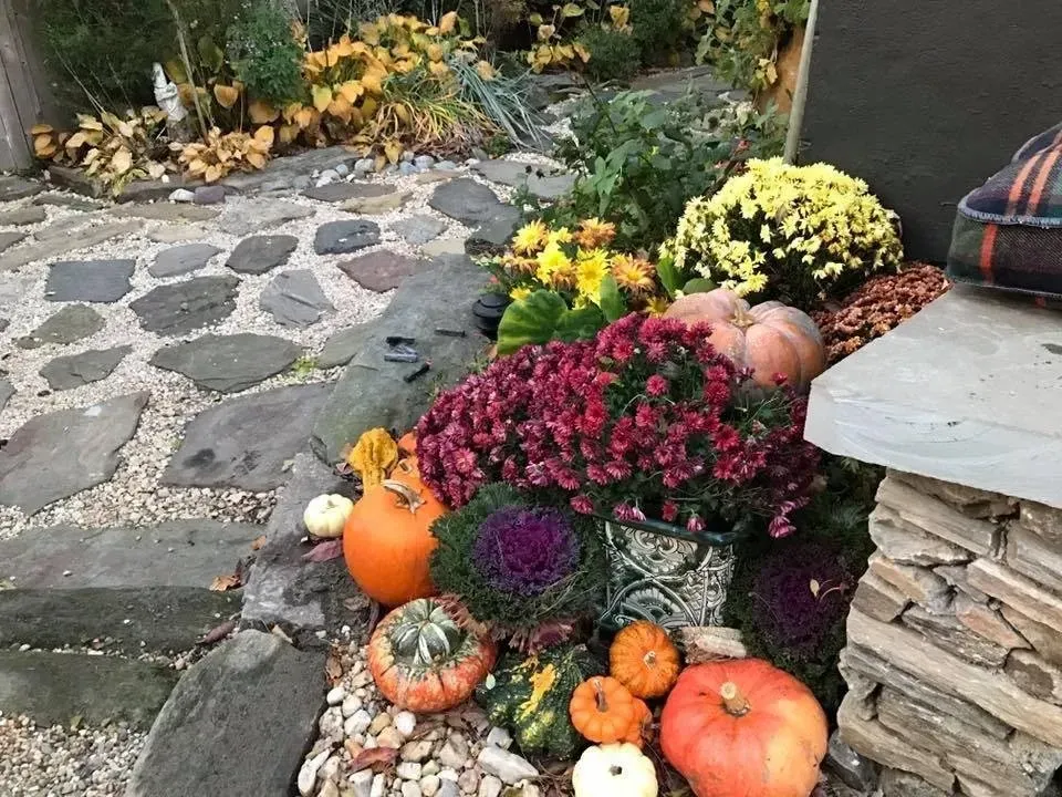 Autumn porch display with pumpkins, chrysanthemums, and stacked stone beside a flagstone path