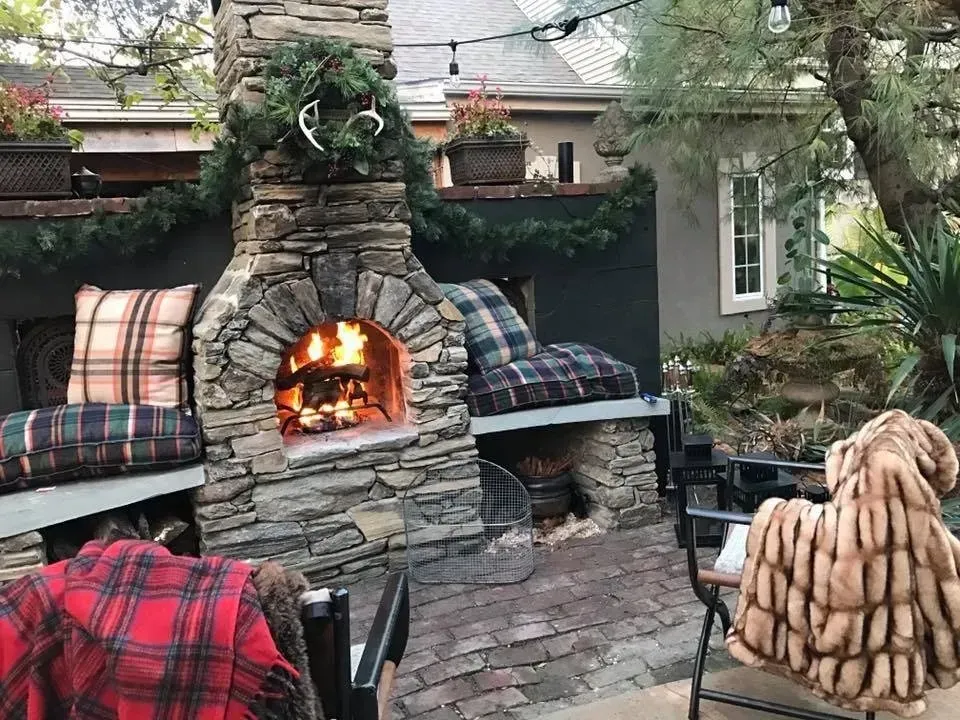 Stone outdoor fireplace on a brick patio with plaid cushions and a cozy fire under trees