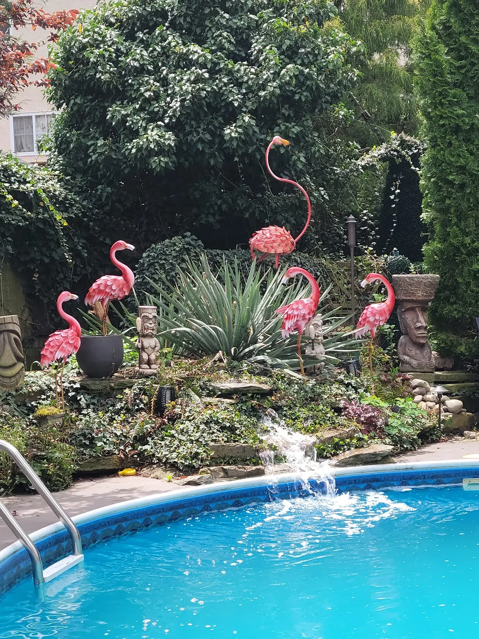 Poolside garden with pink flamingo lawn ornaments among greenery and a blue swimming pool