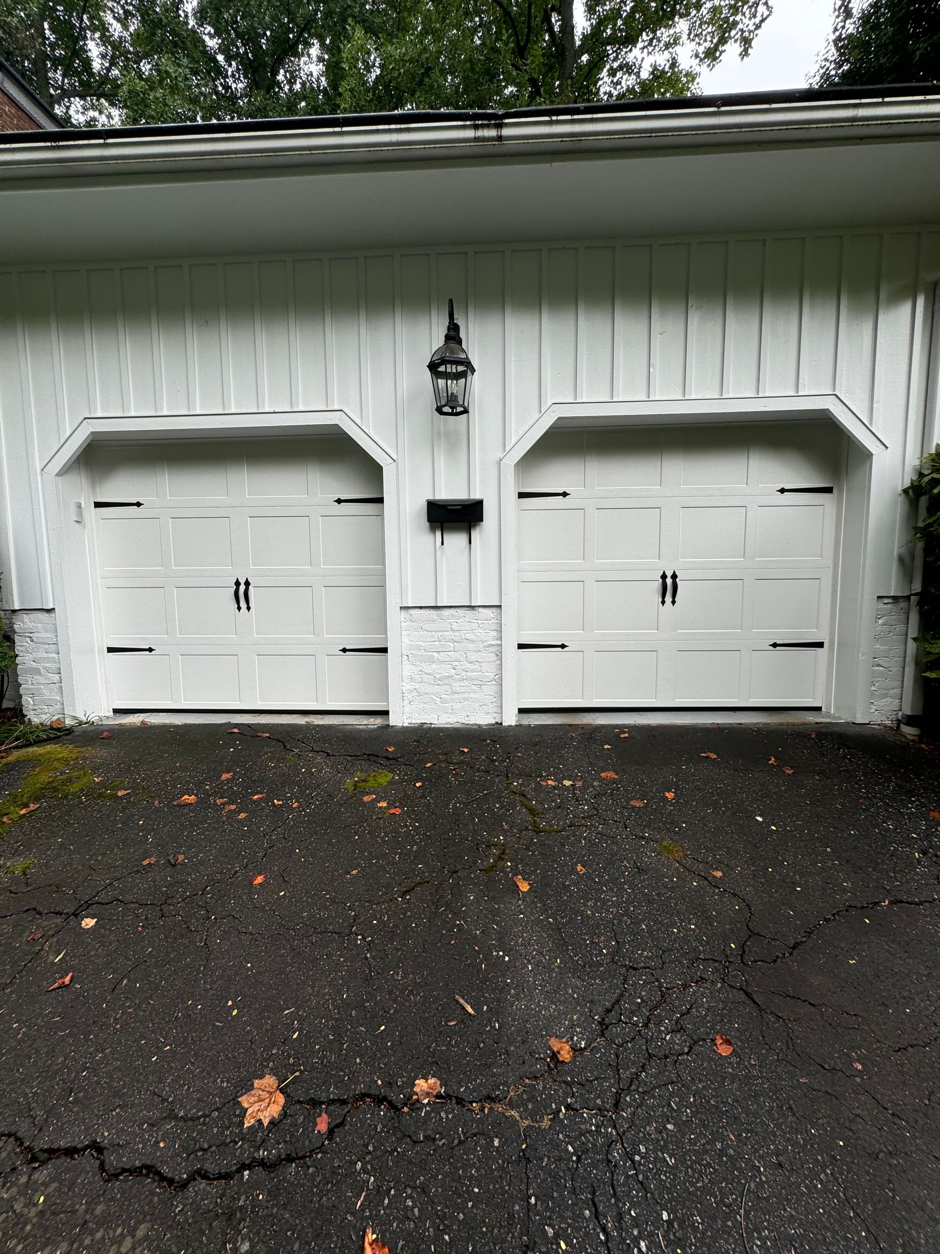 White garage doors with black hardware, on a building with a cracked asphalt driveway.