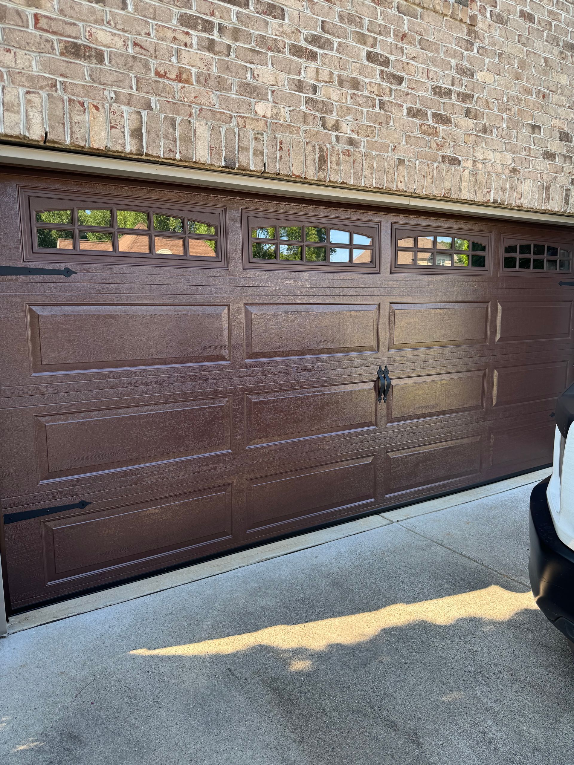Brown garage door with decorative windows, against a brick wall, sunlight on concrete.