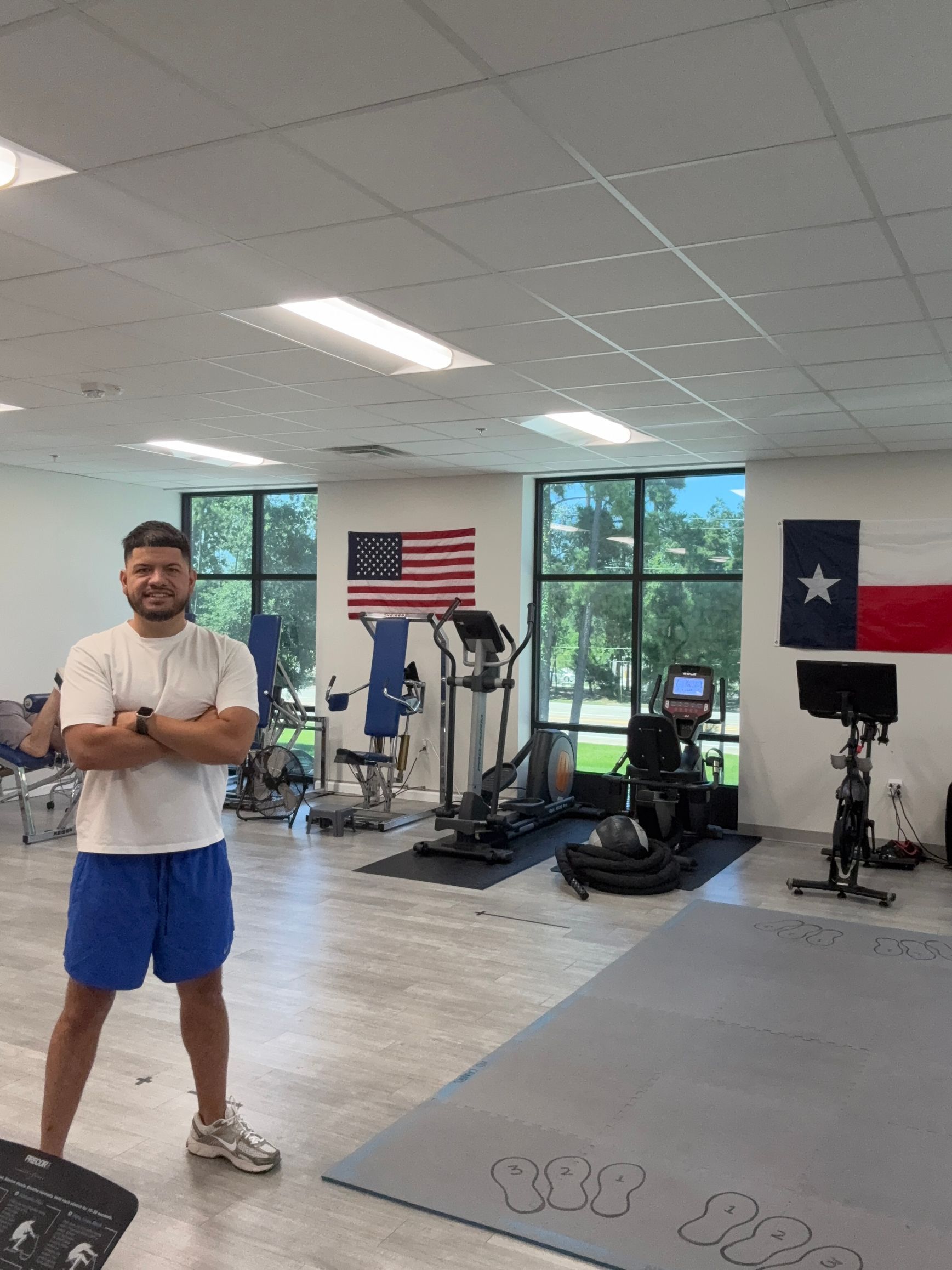 Brandon standing in a gym with exercise equipment, windows, and American and Texas flags in the background.