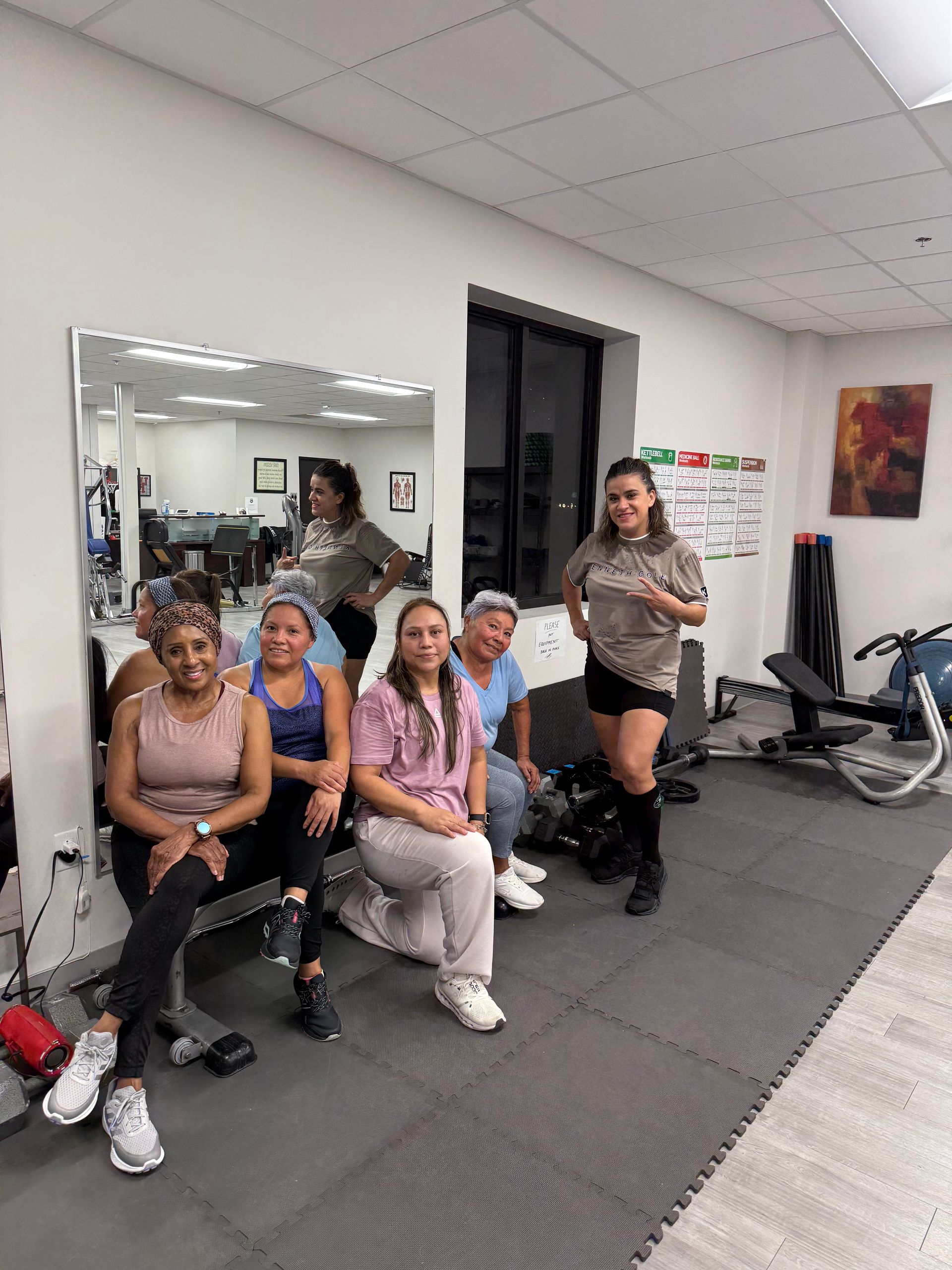 Five women posing in a fitness studio, seated by a wall mirror with exercise equipment nearby.