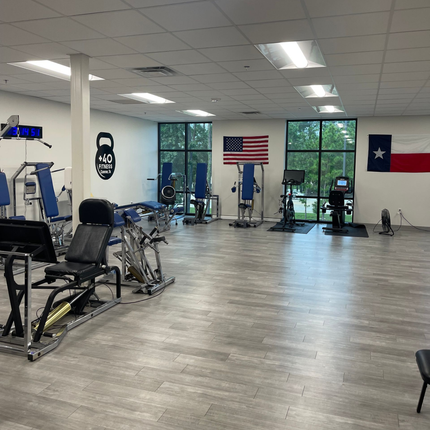 Empty gym with exercise machines, wood floor, Texas and U.S. flags, and large windows.