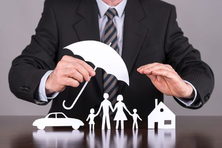A person in a suit holds a paper umbrella over cutouts of a car, family, and house, symbolizing insurance protection.