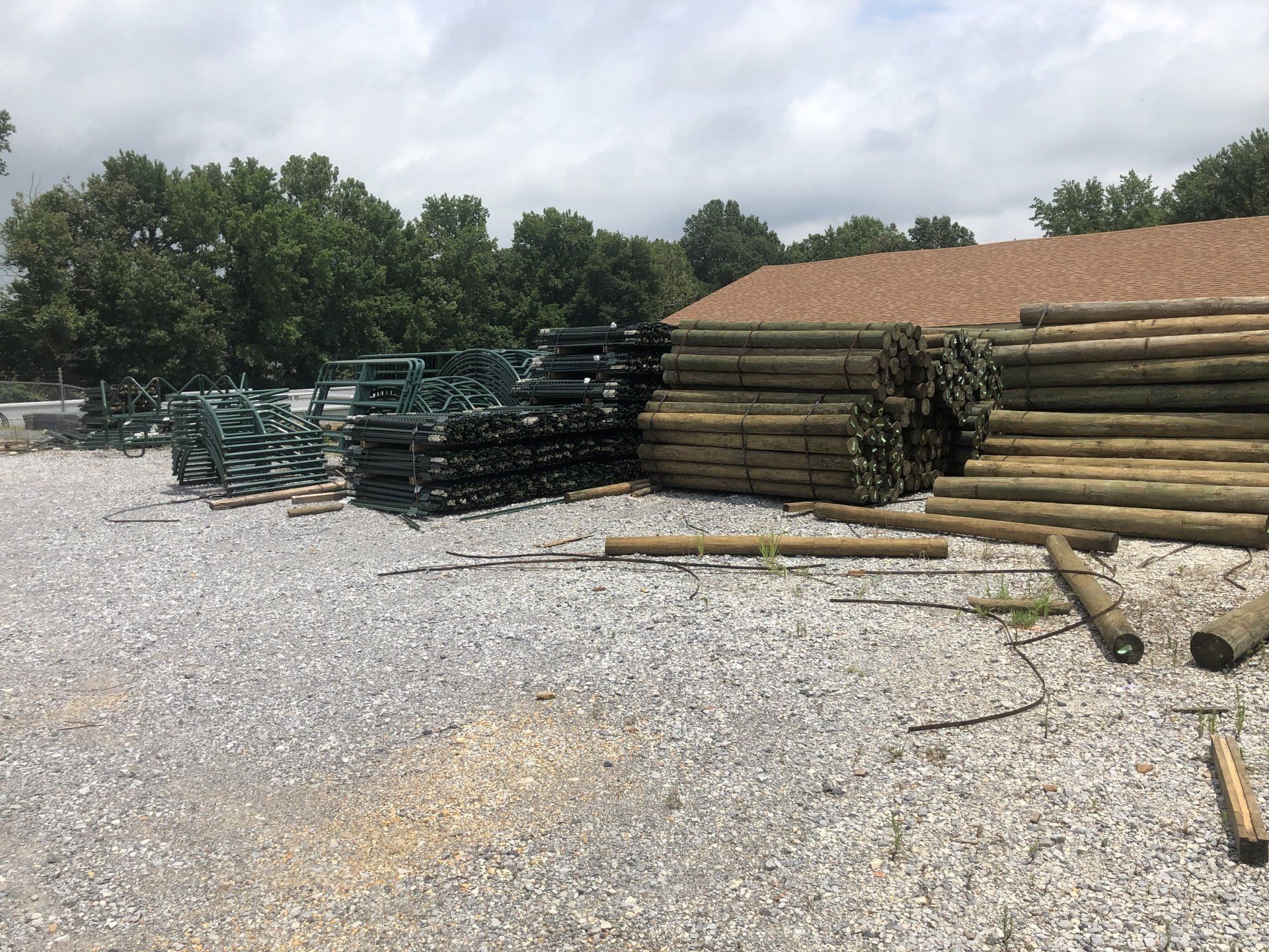 Piles of wooden fence posts and green metal fencing stacked outdoors on gravel, next to a building and trees.