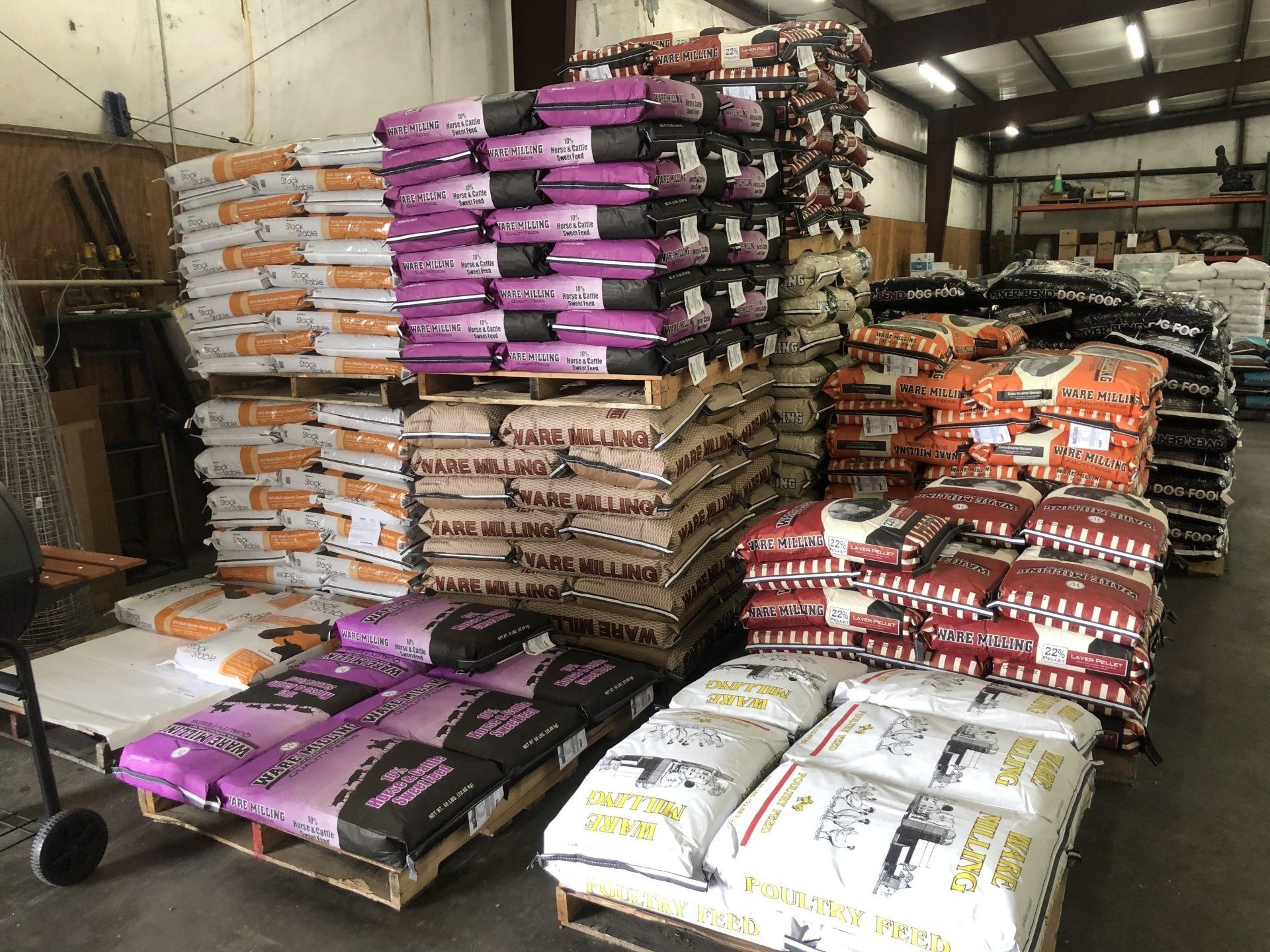 Stacks of feed bags in a warehouse on pallets; bags are various colors including orange, purple, and brown.