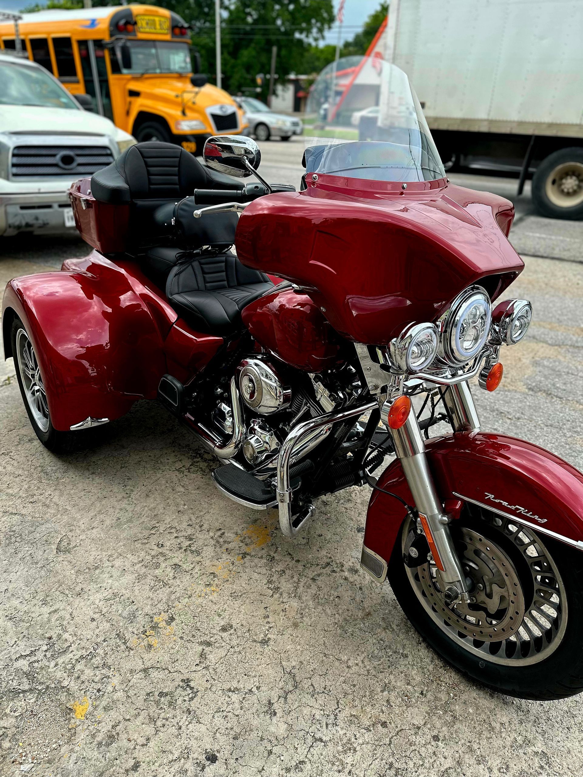 A red motorcycle with three wheels is parked in a parking lot next to a school bus.