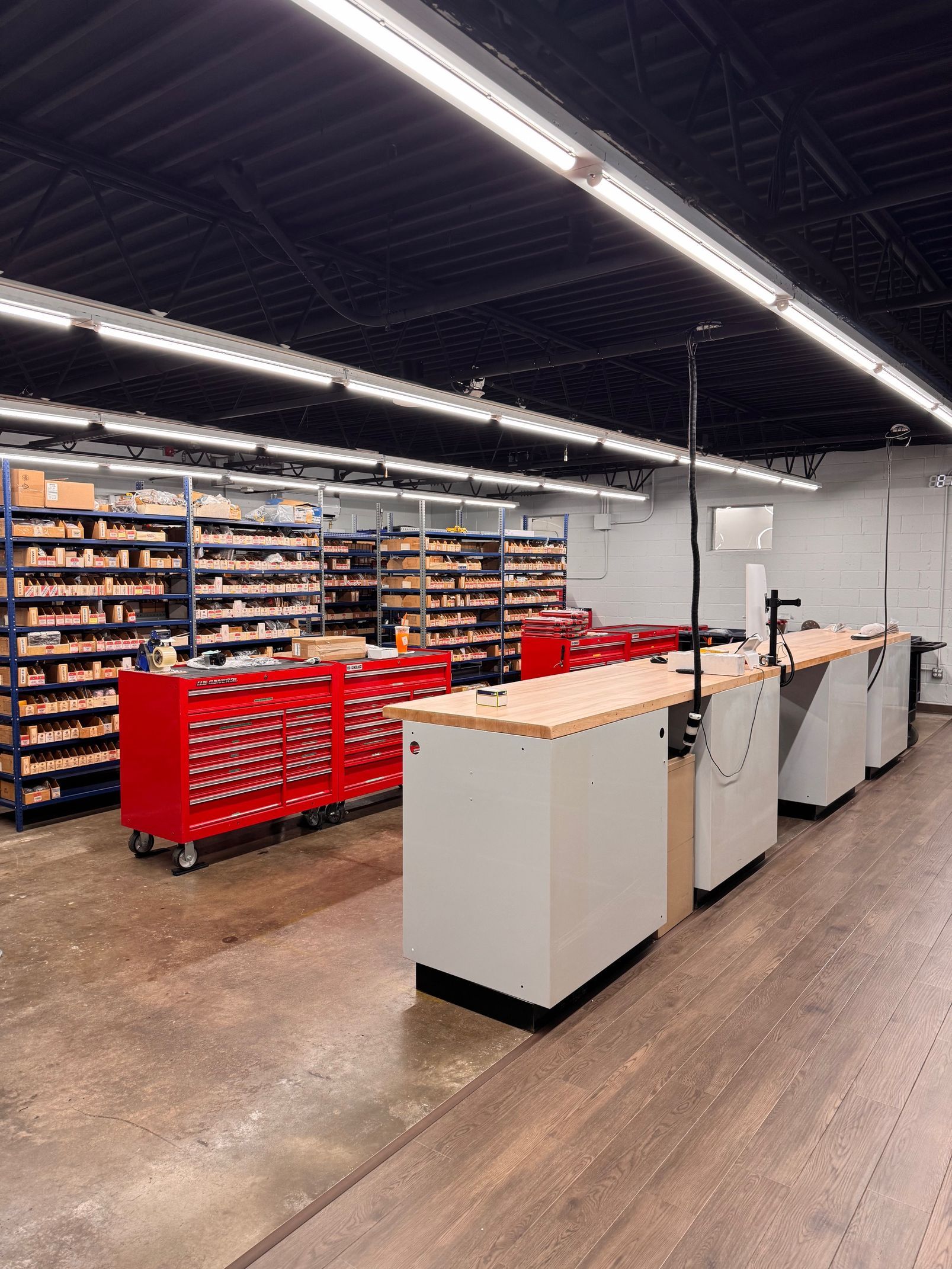 A workshop with red tool chests, wooden workbenches, shelving with parts, and fluorescent lighting.