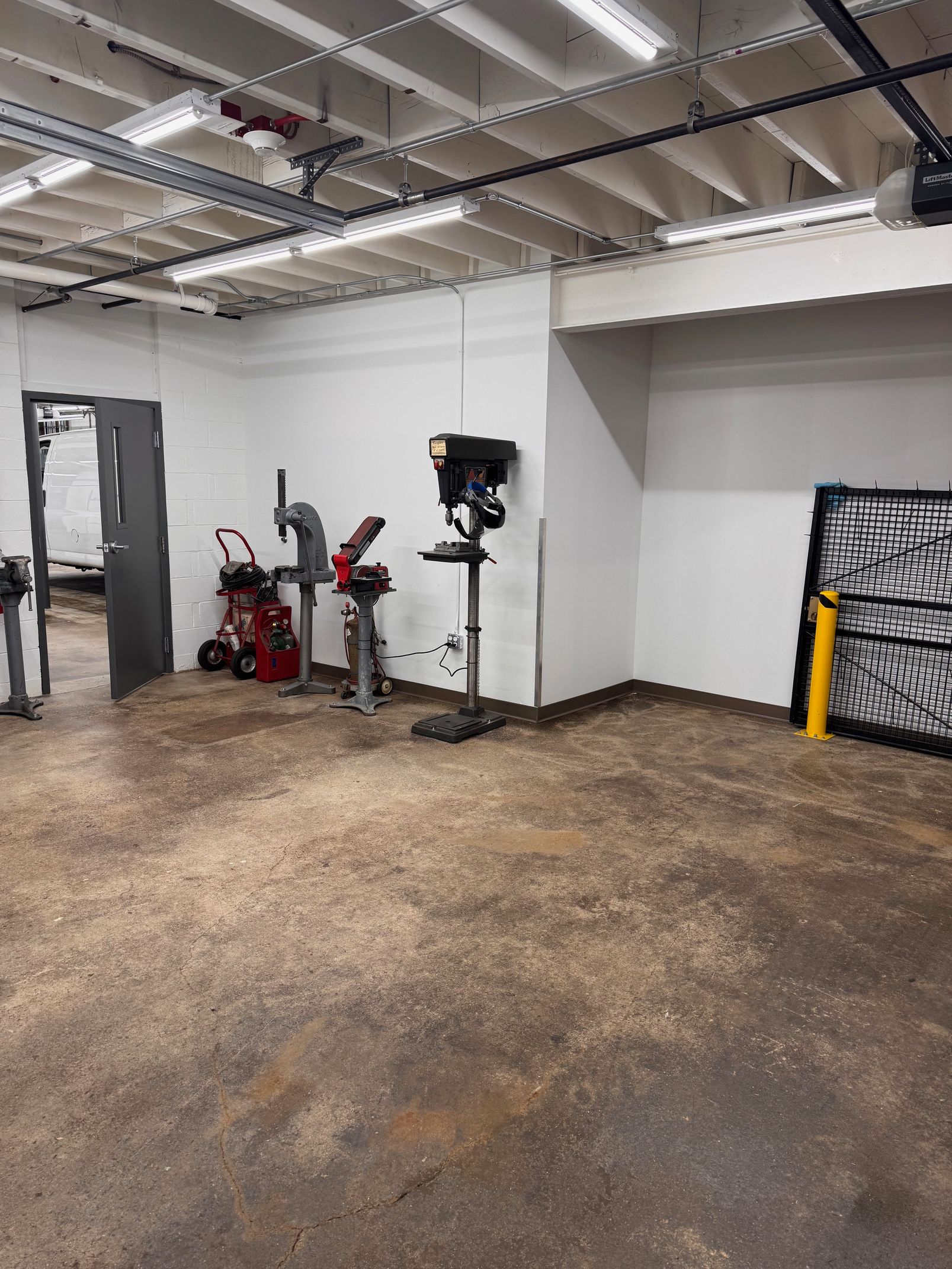 Workshop with drill presses, tools, and sawdust-covered floor, gray door, white walls, bright lights.