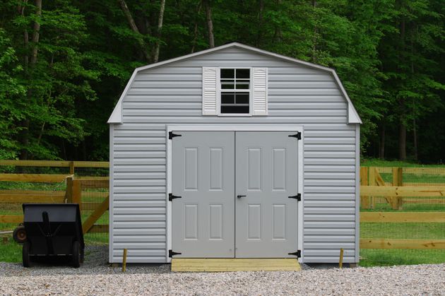 Gray backyard storage shed with double doors and a small window, set on gravel with trees behind it