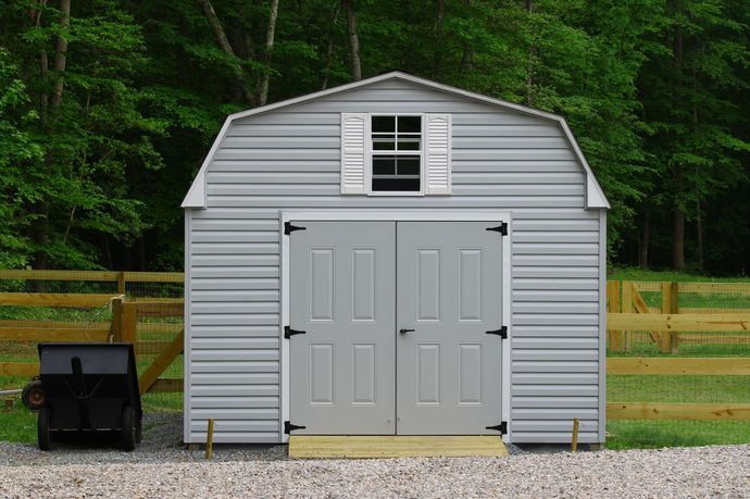 Gray backyard storage shed with double doors and a small window, set on gravel with trees behind it