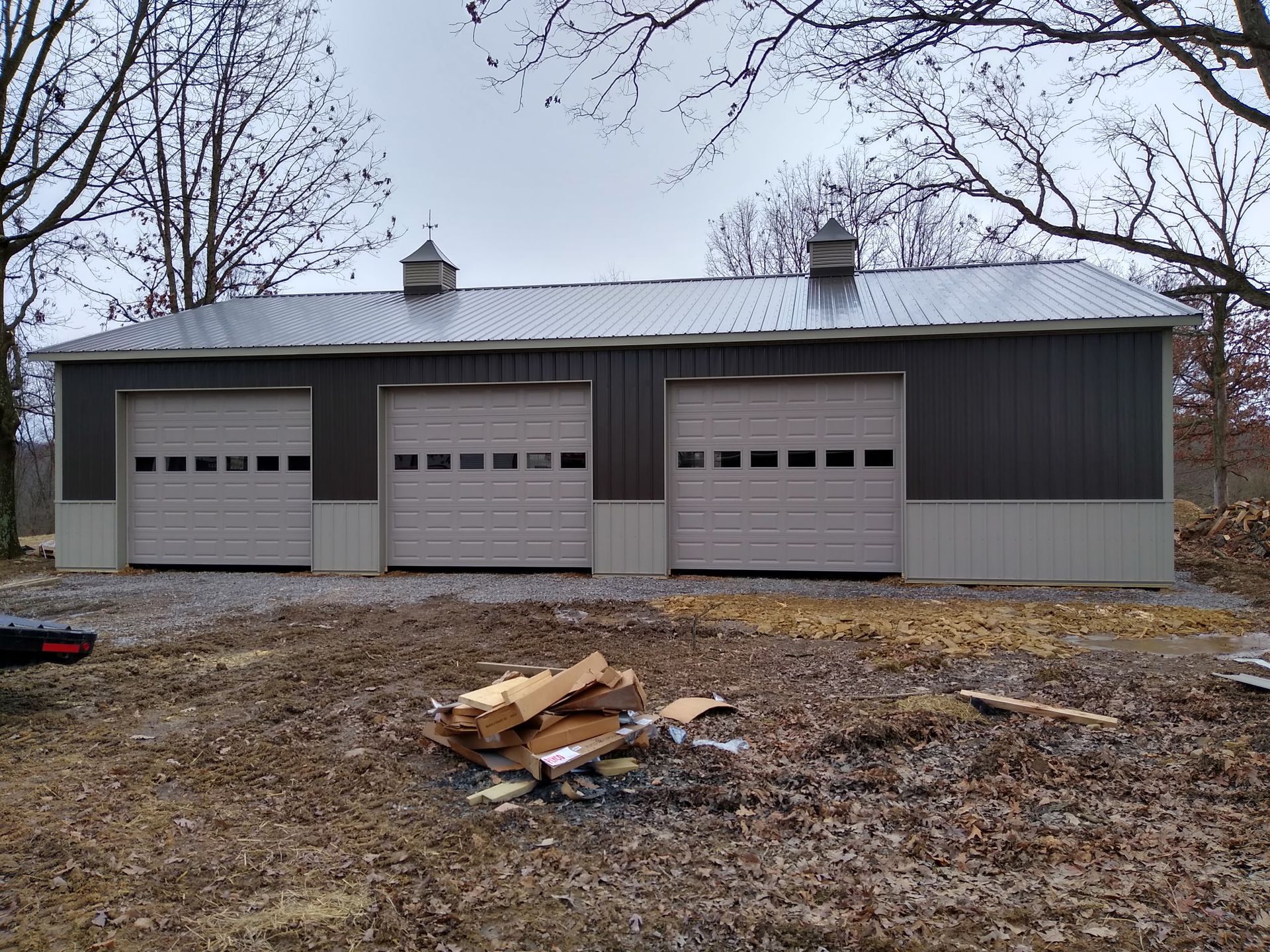Gray three-car garage with white doors in a leafless yard, with wood debris in front.