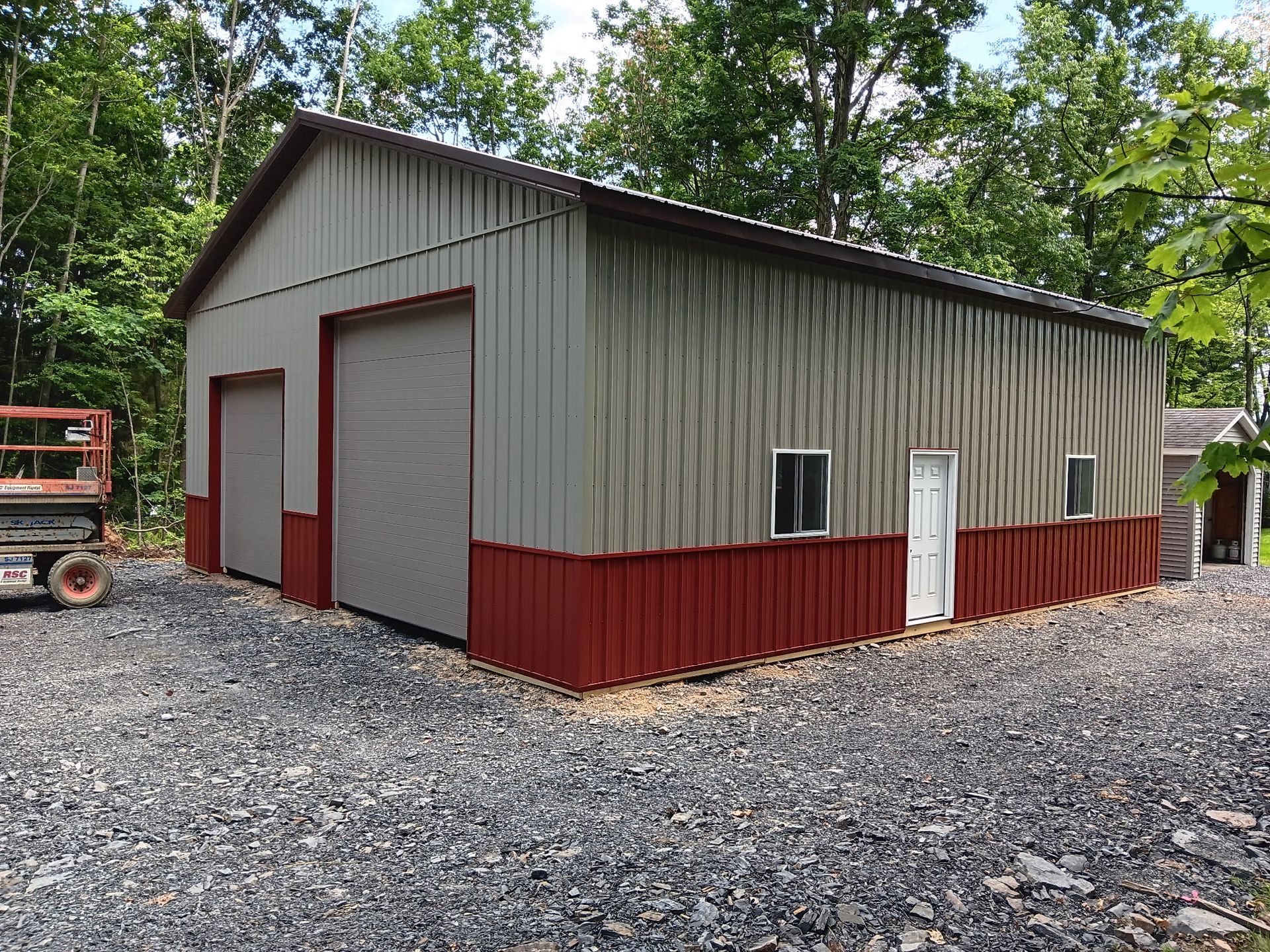 Gray metal barn with red lower siding and doors on a gravel lot, surrounded by trees