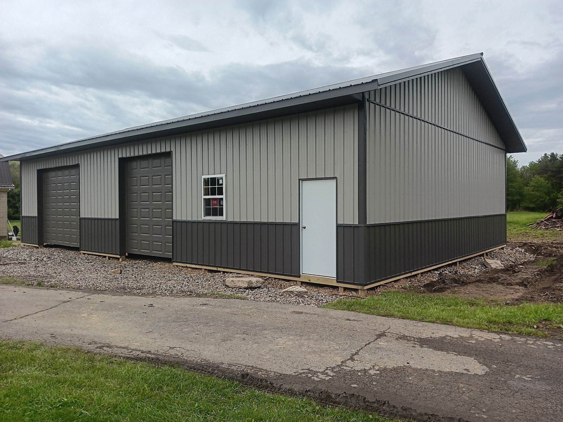 Gray metal warehouse building with garage doors beside a gravel road under an overcast sky