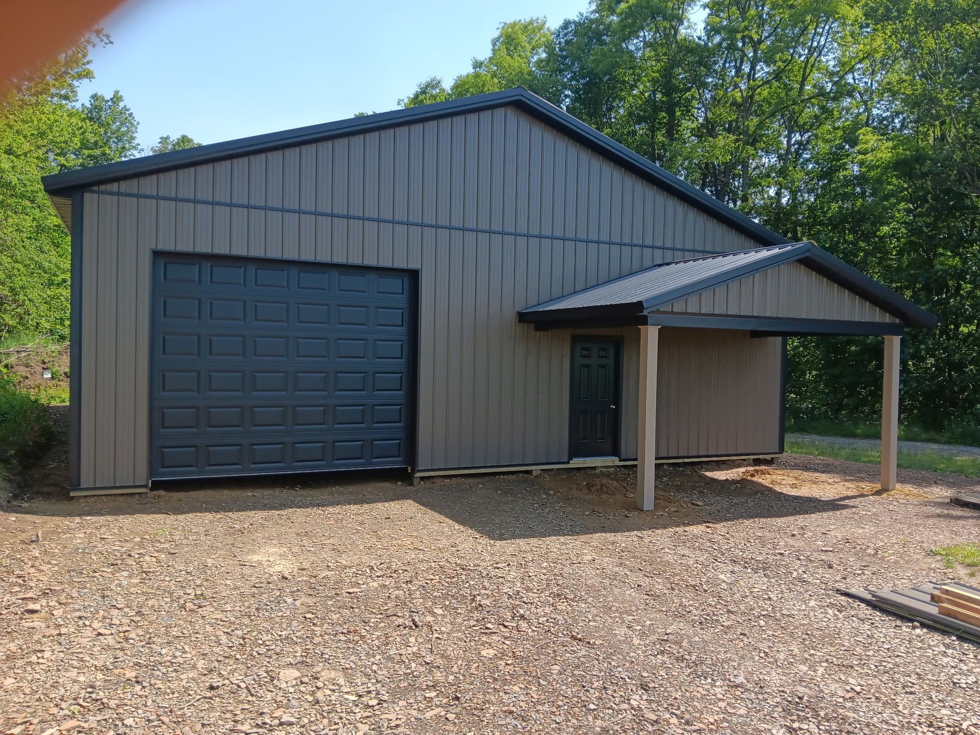 Gray metal garage with a large door and covered entry, set on a gravel lot with trees behind it