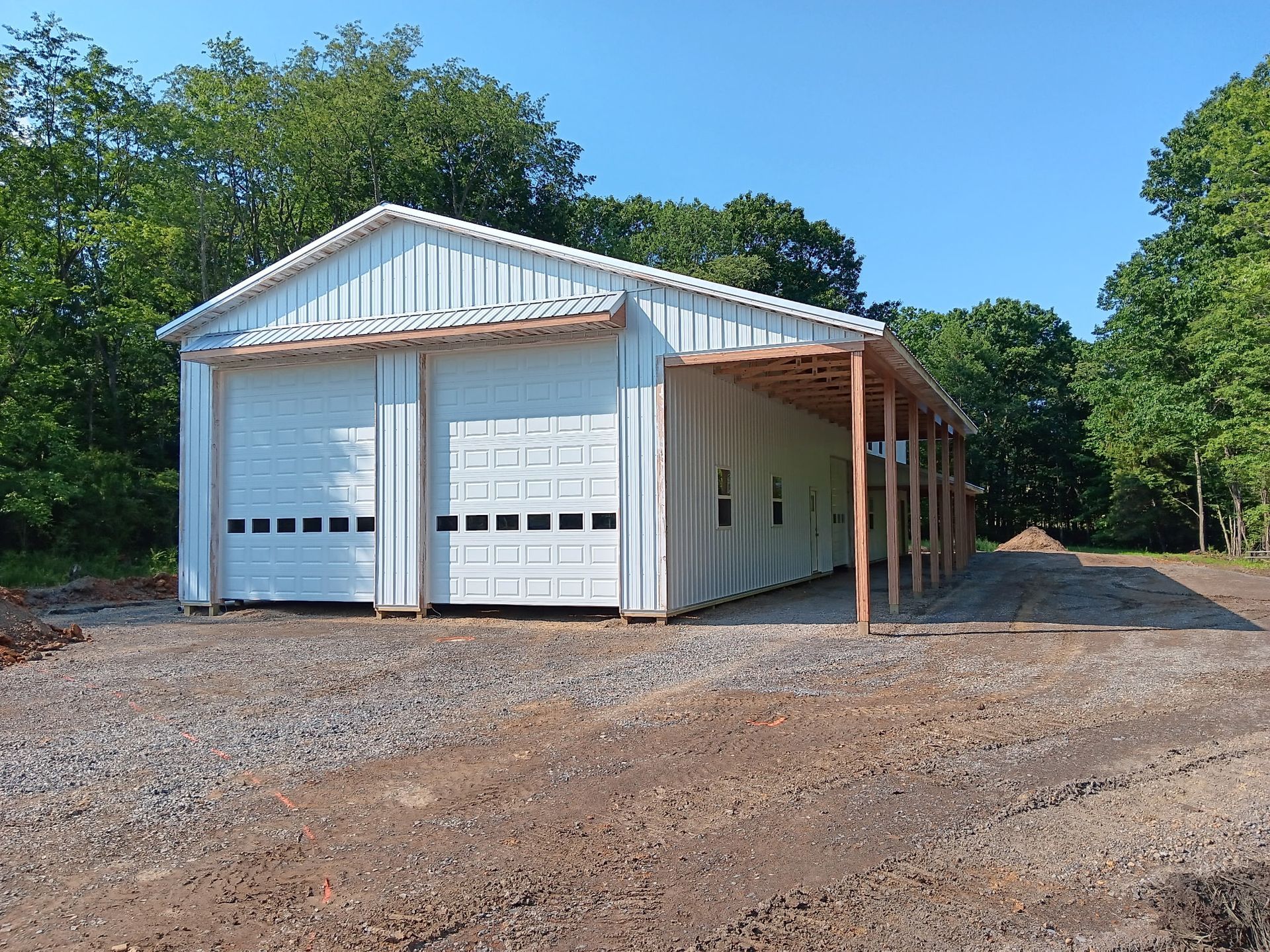 White metal garage/workshop with two roll-up doors and an attached open-sided lean-to on a gravel lot.