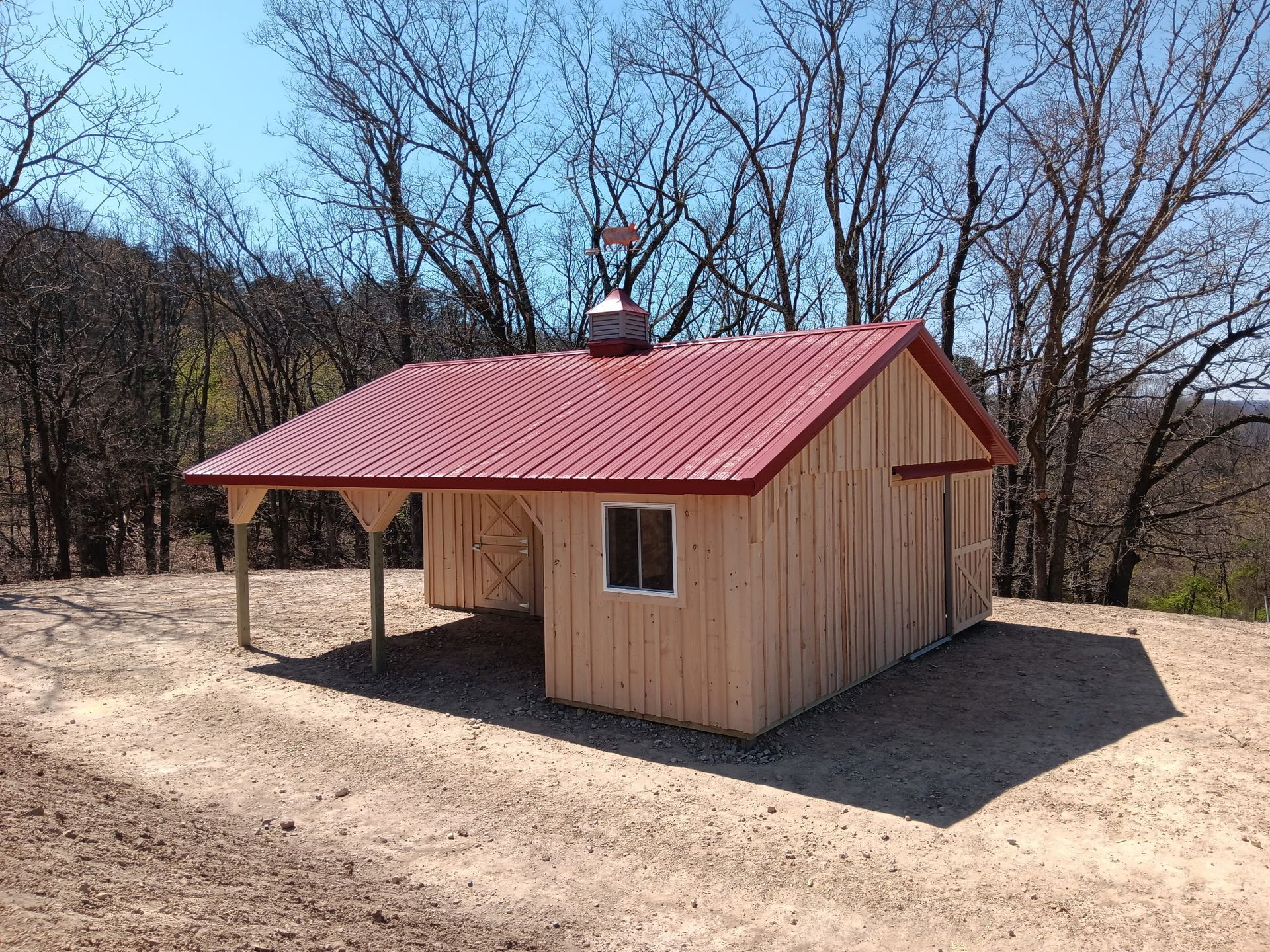 Small wooden cabin with a red metal roof on a dirt clearing, surrounded by bare trees