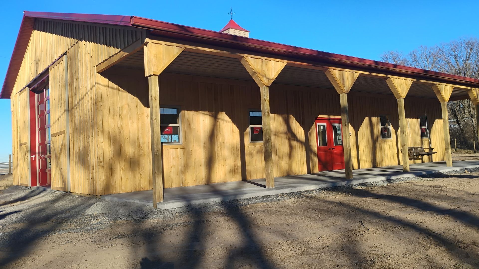 Wooden building with red roof and covered porch in bright sunlight, casting long shadows on gravel.