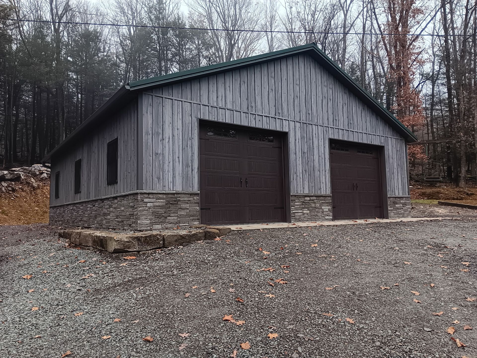 Gray barn with two brown garage doors on a gravel lot in a wooded area