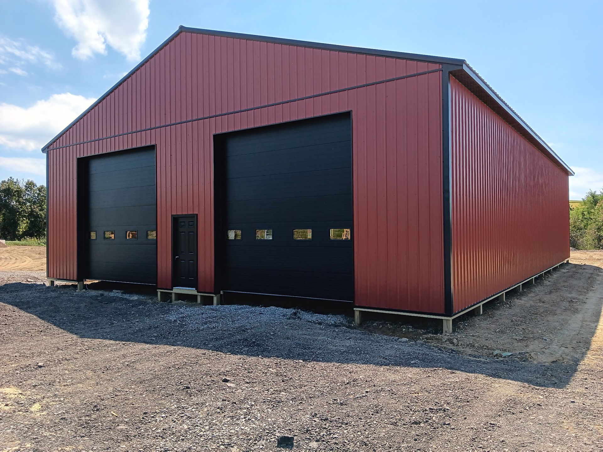 Red metal storage building with two large black garage doors on a gravel lot under a blue sky