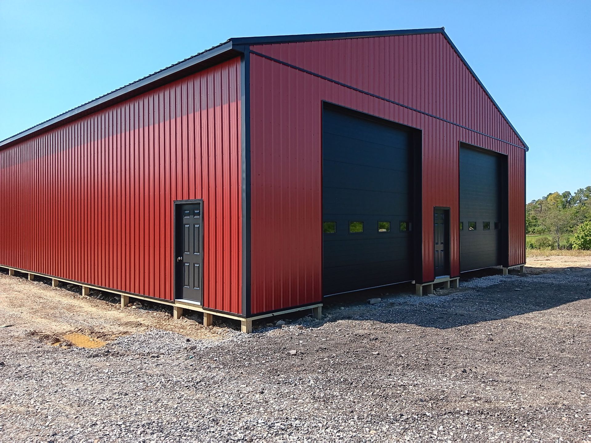 Red metal storage building with black garage doors on a gravel lot under a clear blue sky