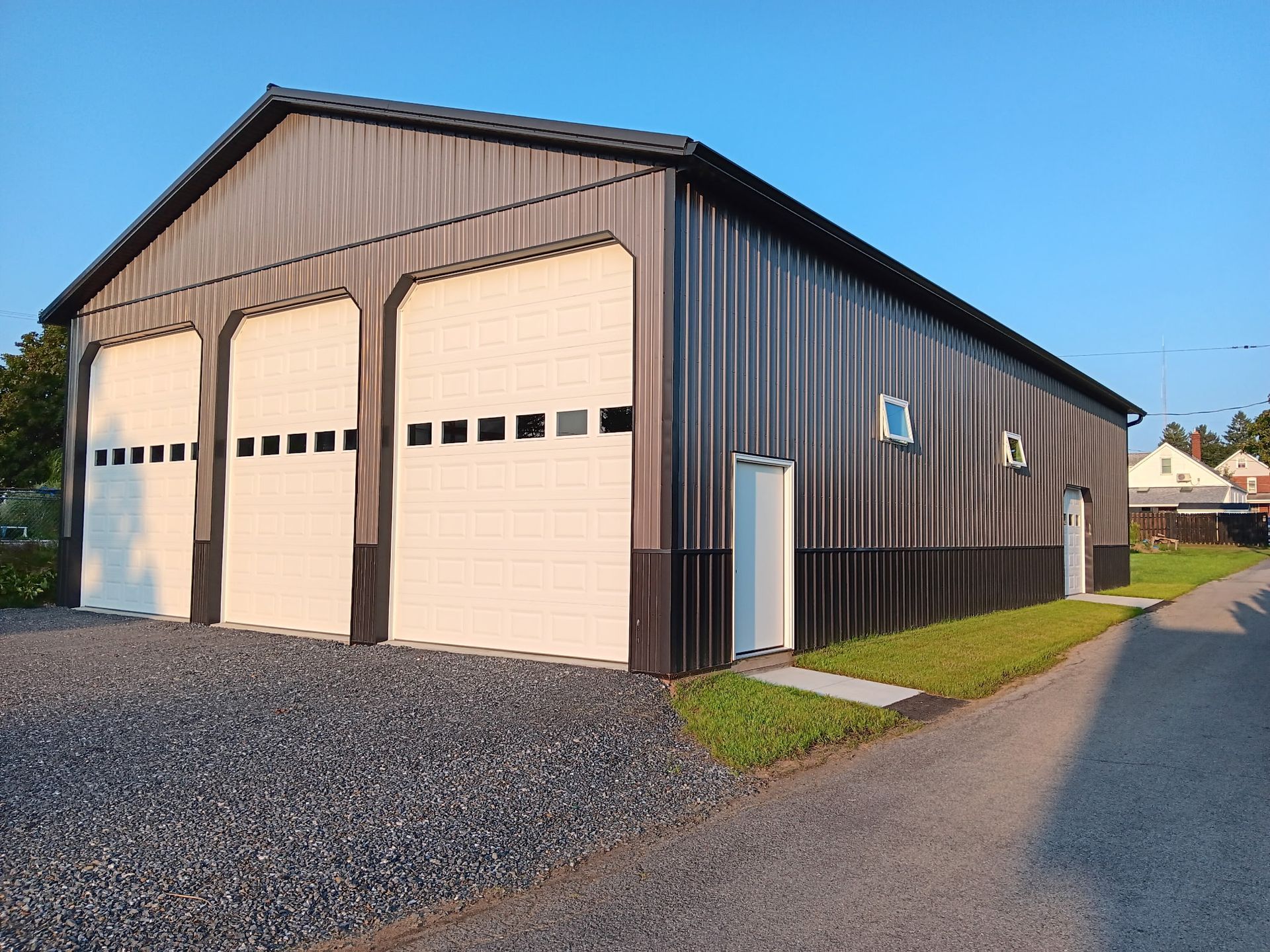 Large brown metal garage with three white roll-up doors on a gravel driveway under a blue sky