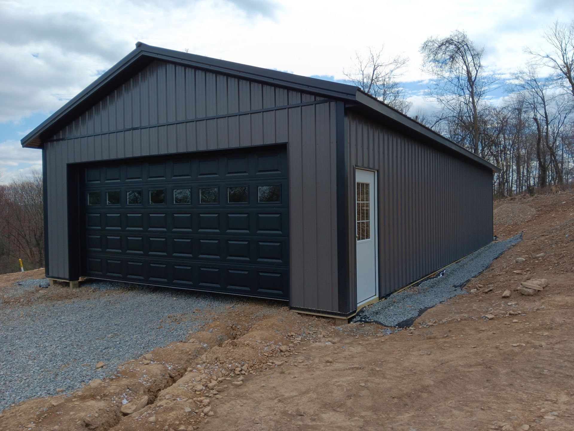 Dark gray detached garage with black door on a gravel drive beside a wooded slope