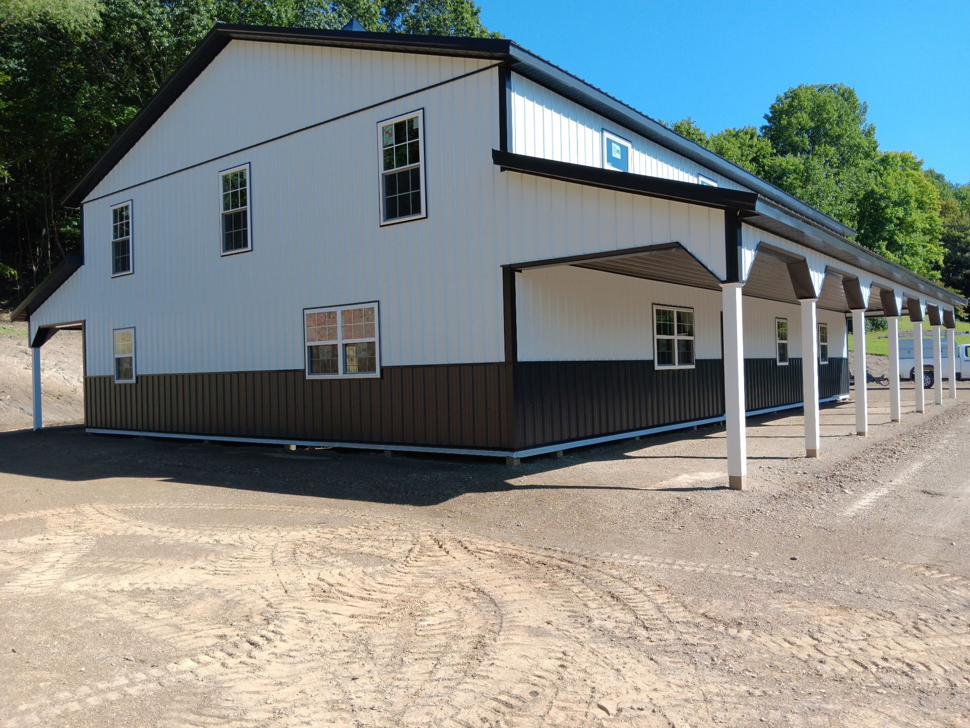 White barn-like building with brown trim and a covered porch on a dirt lot under a blue sky