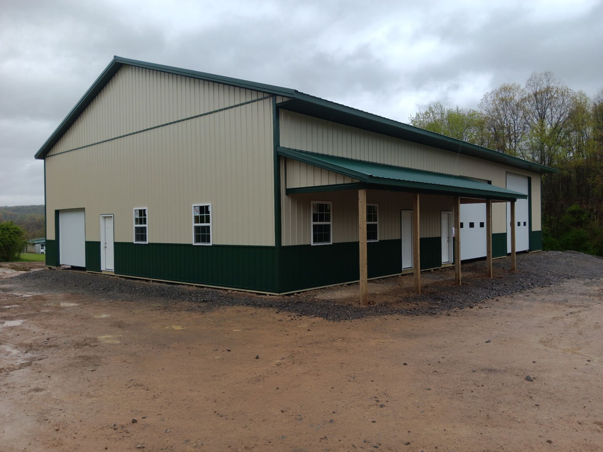 Beige and green metal barn-style building with covered porch on a muddy lot.
