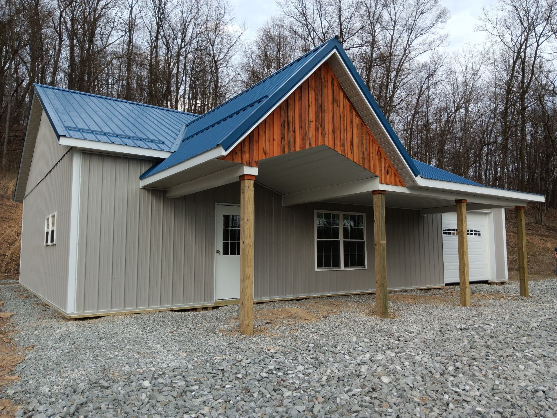 Small gray house with blue metal roof and porch on gravel, surrounded by leafless trees
