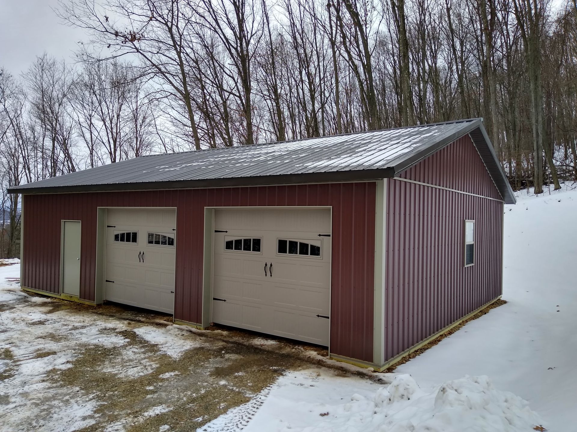 Two-bay garage with maroon siding and white doors in a snowy yard with bare trees.