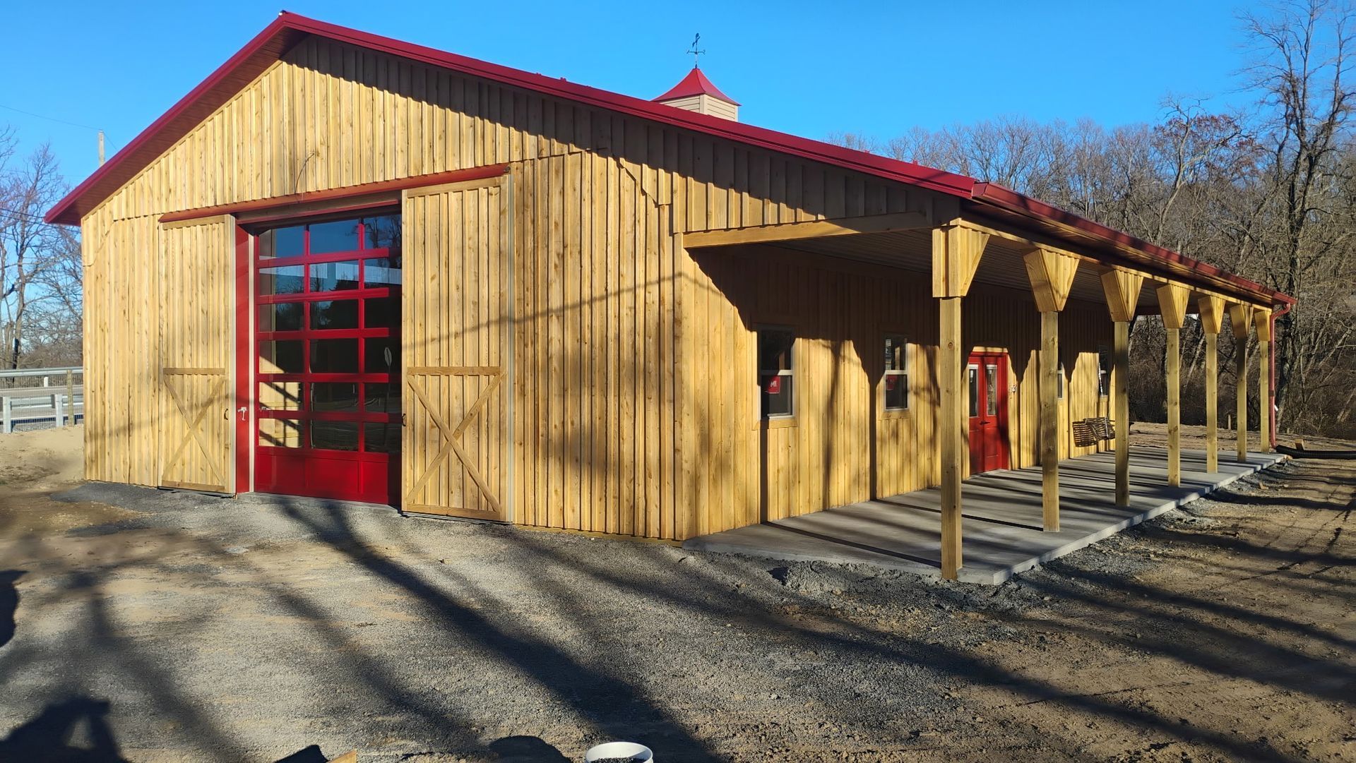 Wooden barn with red trim and roof, set in a gravel lot under a clear blue sky.