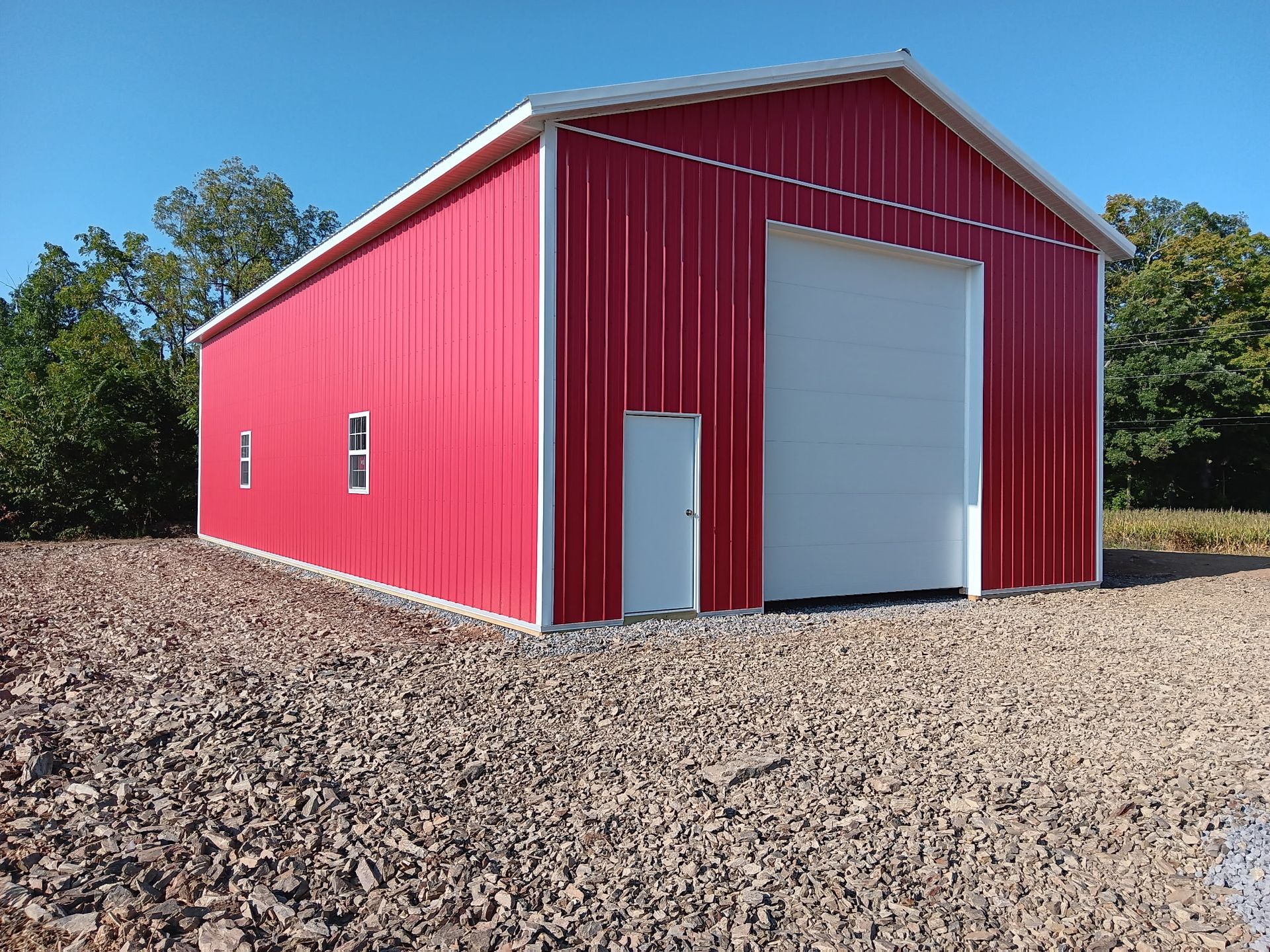 Red metal storage building with white garage door on a gravel lot, surrounded by trees.