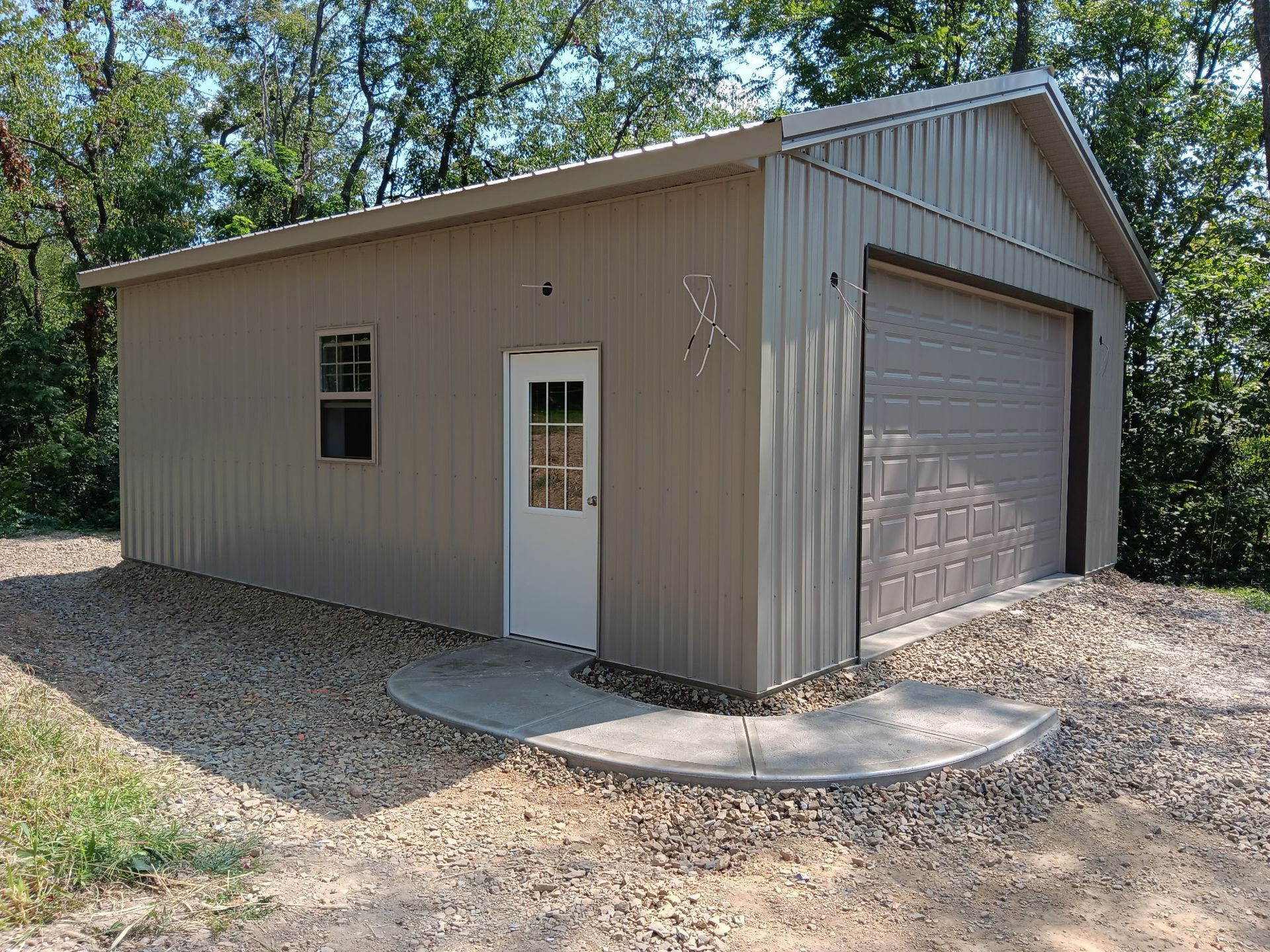 Tan metal garage-style shed with white door and brown roll-up door on a gravel lot among trees
