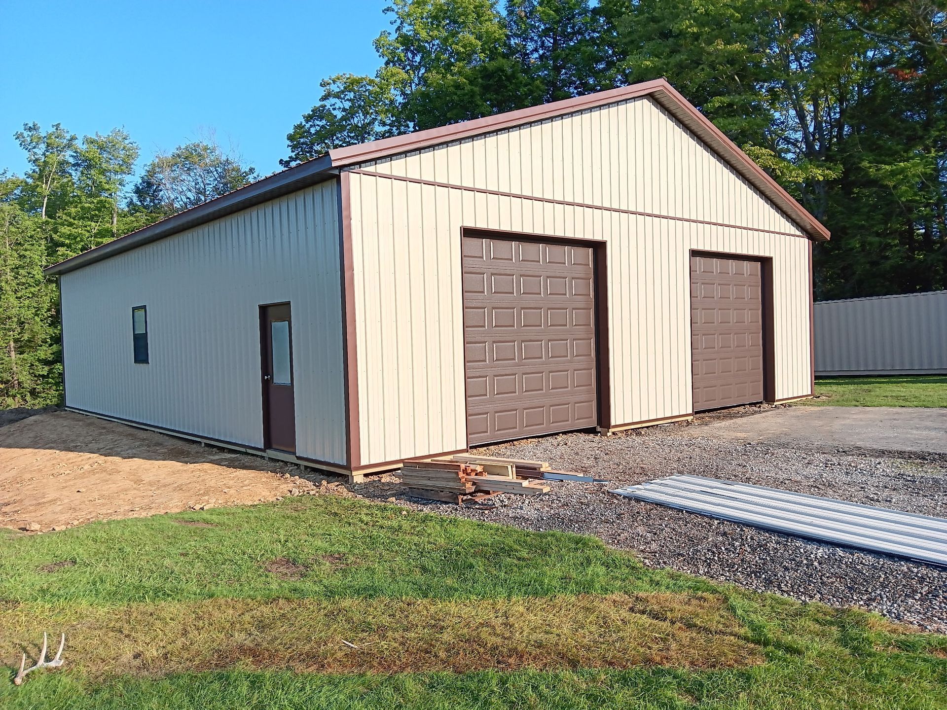 Beige metal garage with two brown doors on a gravel driveway, surrounded by grass and trees.