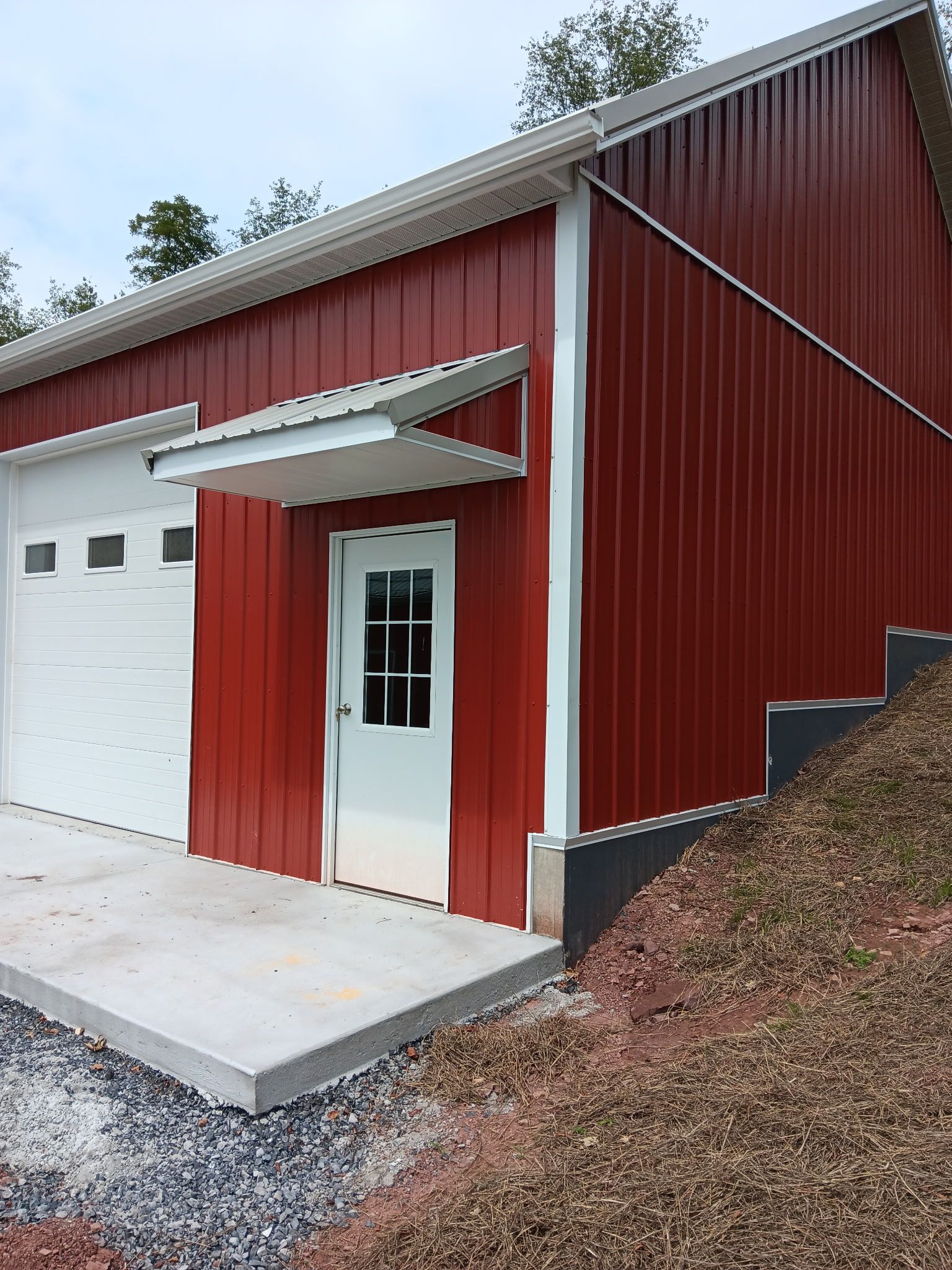 Red metal-sided garage with white door and concrete driveway on a wooded slope