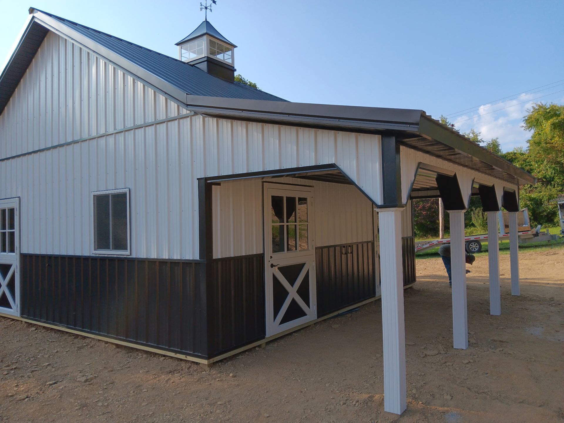 White barn-like building with black lower siding and a covered porch on a dirt lot under a blue sky