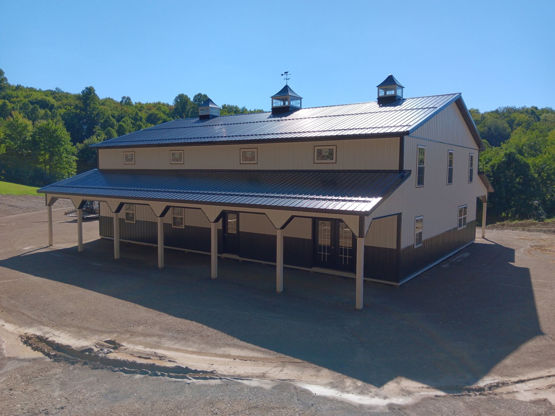 White barn with black roof and cupolas on a gravel lot, surrounded by trees under a clear sky