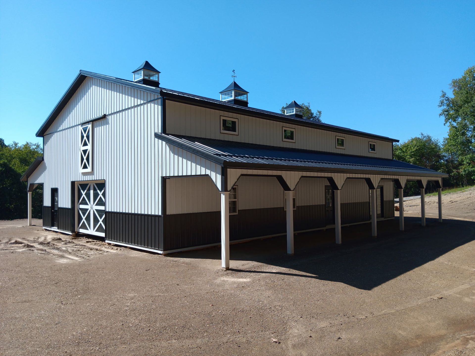 White barn with black trim and covered side porch on a dirt lot under a clear blue sky