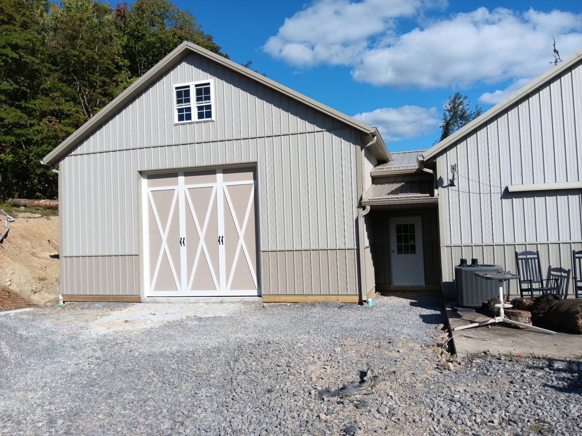 Gray barn with white X doors beside a narrow walkway and a larger white building on gravel under a blue sky