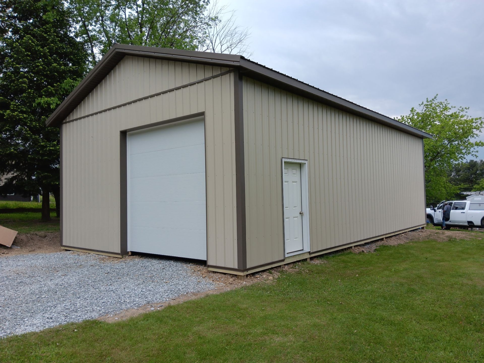 Tan metal storage building with roll-up garage door, side entry door, and gravel driveway.